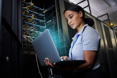 Laptop, network and data center with a black woman it support engineer working in a dark server room. Computer, cybersecurity and analytics with a female programmer problem solving or troubleshooting.